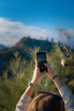 A girl with blond hair takes pictures of nature on the phone. You can see mountains, sky, clouds and coniferous tree
