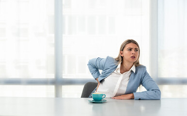 Young business woman having back pain ache from bad posture while sitting at desk in office on non ergonomic chair at the work place in formal wear with cup of coffee