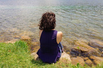 Fototapeta na wymiar Back view of a young woman sitting near a lake. Concept of relaxation, nature, envinronment. Copy space.