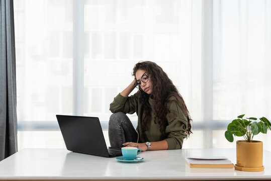 Stressed young woman holding head in hands and feeling demotivated while sitting at her home office and working remotely on laptop. Depressed female student tired of online learning and education