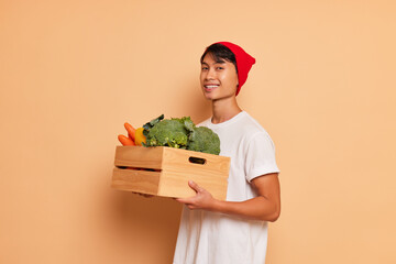 Cheerful Asian guy in white t-shirt and red hat standing inside the studio holding box of fresh...