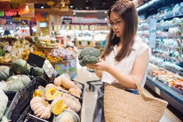 an asian woman holding for shopping a piece of broccoli in a grocery store, vegan lifestyle concept.