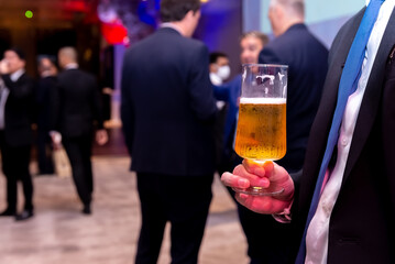 A man in a suit stands at a feast, holding a glass of beer. He is surrounded by other people, all of whom are also drinking and eating. The atmosphere is festive and relaxed.