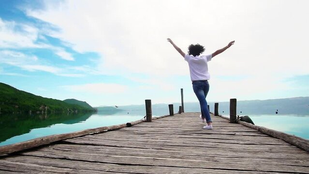 Positive traveler woman show victory, success emotions running and jumping on wooden jetty pier with hands wide outstreatched, slow motion