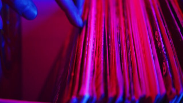 DJ carefully flipping through the records in the box to choose the right one to use in his DJ set during the studio recording of the vinyl session in a color light close up