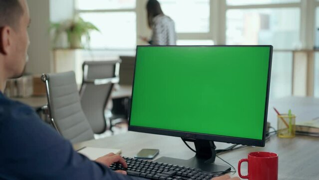 Over the Shoulder: Young Man Sitting at His Desk Uses Desktop Computer with Green Mock-up Screen.Professional Office Employee Working on Computer