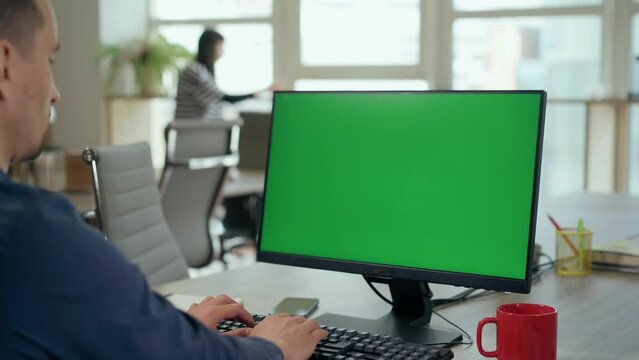 Over the Shoulder: Young Man Sitting at His Desk Uses Desktop Computer with Green Mock-up Screen.Professional Office Employee Working on Computer