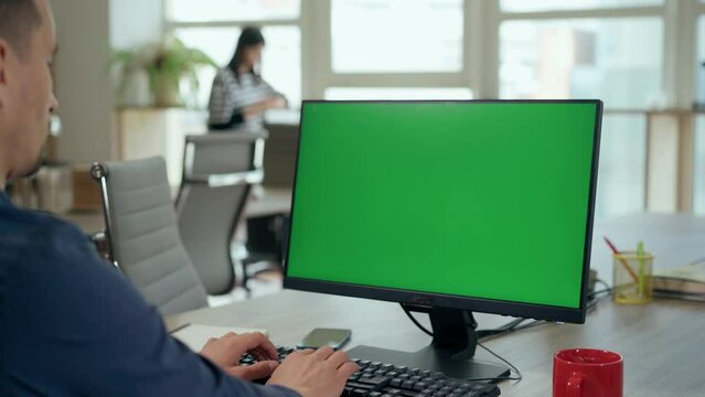 Over the Shoulder: Young Man Sitting at His Desk Uses Desktop Computer with Green Mock-up Screen.Professional Office Employee Working on Computer