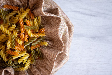 Uncooked multicolored Fusilli pasta in fabric bag over white wood background. top view.
