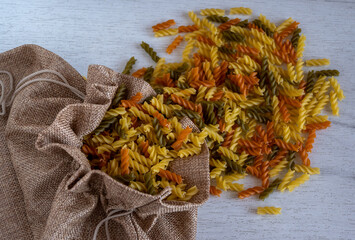 Uncooked multicolored Fusilli pasta spilled out of fabric bag over white wood background. top view.