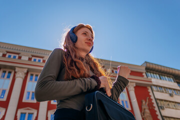 Smiley ginger girl with headphones on looking away while holding a lap top case. Low angle view