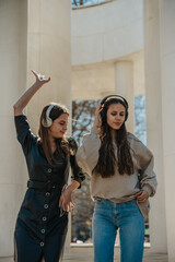 Front view shot of two brunette girls listening to music on their headphones and dancing. Gorgeous girl in black overcoat dancing with one hand up