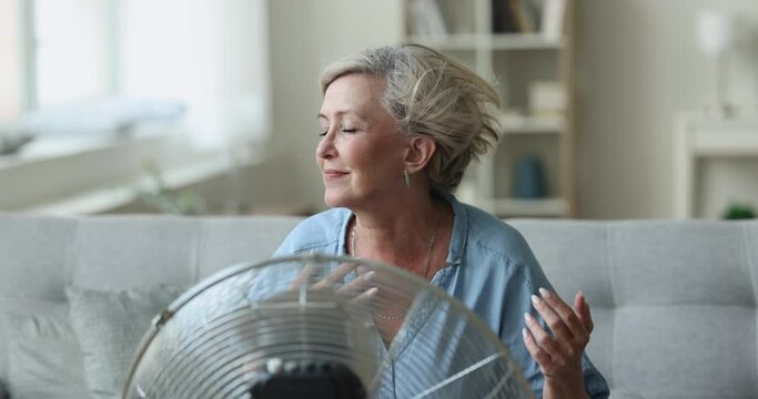 Happy relaxed senior lady sitting at fresh cool air blowing from fan, resting on home sofa, enjoying freshness from electric blower, ventilator, cooler, using home appliance, equipment for ventilation