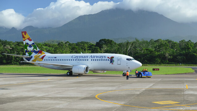 Close view of Cayman Airways airplane on a runway after landing at La Ceiba Airport in Honduras. Mountains with clouds and tropical trees in the background. Travel concept.