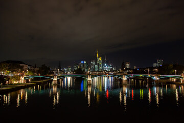 Fototapeta na wymiar Cityscape in Frankfurt, Germany featuring a bridge, river, nighttime city illumination reflection, and a downtown skyscrapers background.
