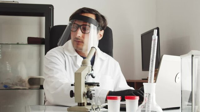 A man in a white coat works on a laptop in a laboratory. The laboratory assistant brings four petri dishes with samples