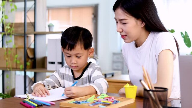 Young beautiful Asian single mother watching her son draw while mother is working from home laptop, adorable boy painting on paper with color pens. Happy family spend time together on holidays.