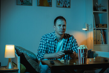 Concentrated caucasian man playing video game on a big screen and eating popcorn at home. Man holding wireless controller of a gaming console in a room filled with blue screen light at night.