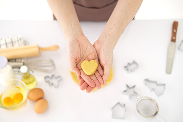 Cooking with love. Female hands holding dough in heart shape. Baking ingredients on the white table in home kitchen. top view
