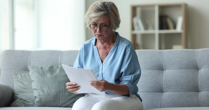 Serious old senior woman in glasses reviewing document, reading paper letter, insurance agreement at home, getting confused, receiving concerning news, medical examination result