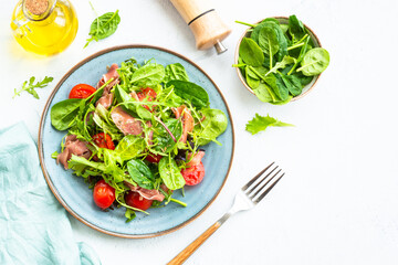 Fresh salad with green leaves, tomatoes and jamon at white table. Top view with copy space.