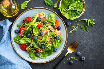 Salad with fresh leaves, tomatoes and jamon at black table. Top view with copy space.