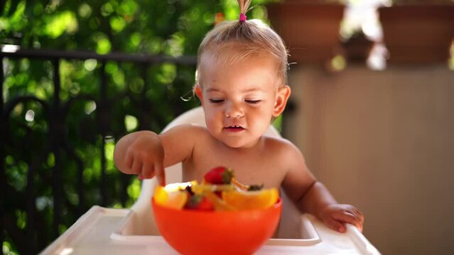 Little laughing girl sitting on a highchair and touching fruits in a bowl