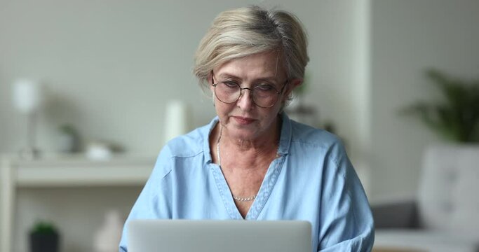 Thoughtful positive elder business woman in glasses working at laptop at home, typing, looking away, thinking on creative ideas, turning to screen, typing, writing article
