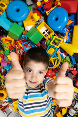 Fototapeta na wymiar Happy 5 year old boy showing thumbs up lying among toys, top view