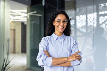Portrait of happy and successful business woman, boss in shirt smiling and looking at camera inside office with crossed arms, Hispanic woman with curly hair in corridor.