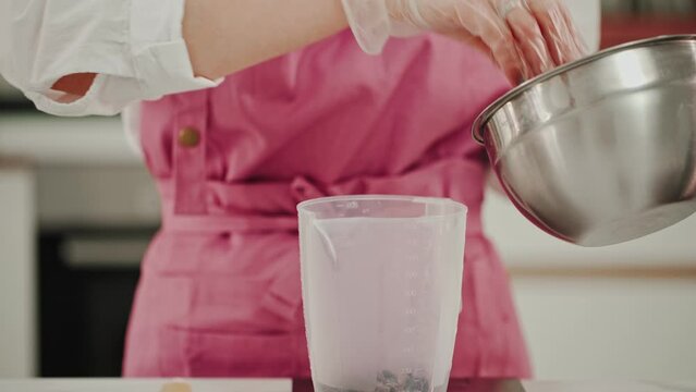 A female confectioner in a pink apron prepares a dessert in a pastry shop. She weighs the required amount of chocolate according to the recipe into a plastic container.