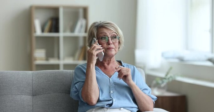 Serious concerned senior woman in glasses talking on mobile phone, speaking to client, doctor, support service hotline operator, telling problem, listening, making call from home couch