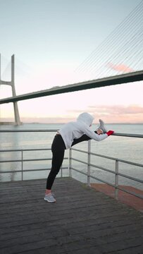 Middle age woman stretching legs before kickboxing exercises. Caucasian athletic female does lunges exercises stretch leg seaside view with bridge and blue sky background. Vertical footage