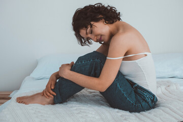 Obraz premium A woman with curly hair sits on a bed with her legs clasped and her eyes closed. The girl hugged herself. 