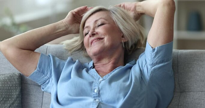 Peaceful happy elderly woman resting on home sofa, leaning on back, stretching body, breathing fresh air, opening closed eyes, looking away, smiling, enjoying leisure, relaxation
