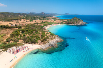 Aerial view of white sandy beach, umbrellas, mountains, green trees, yacht in blue sea at sunset in summer. Tropical landscape. Travel in Sardinia, Italy. Top view of ocean with transparent water