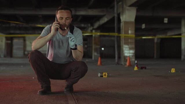 Full shot of young Caucasian male police detective or FBI agent in plain clothes squatting on ground at crime scene, examining cigarette stub, talking on smartphone, and red, blue car lights flashing