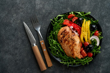 Salad with grilled chicken breas fillet with fresh arugula, tomatoes and olives on black plate on old dark background. Chicken meat with salad. Ketogenic lunch. Healthy food. Top view.