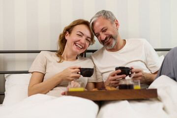 Mature couple enjoying coffee and breakfast at home in bed