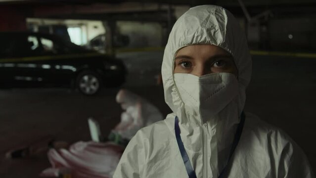 Medium close-up portrait shot of female forensic specialist in protective suit and face mask looking at camera with shock and stress, while colleague is collecting samples off corpse at crime scene