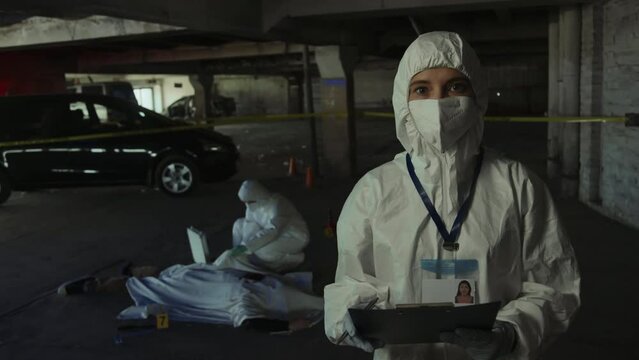 Medium portrait shot of woman in white protective suit and face mask standing at murder scene, writing on clipboard, then looking at camera, and colleague examining corpse of victim in background