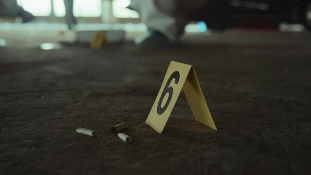 Close-up shot of yellow police marker with number 6 on ground next to cigarette stubs at urban crime scene, and unrecognizable forensic experts in white suits collecting evidence in background