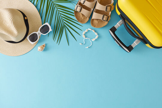Concept of summertime travel. Top view flat lay of yellow suitcase beach accessories and palm leaf on light blue background with empty area for text or promotion