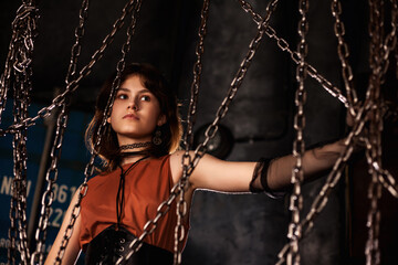 Graceful teen girl model 15-16 year posing in dark industrial room, looking away through chains. Cute classy teenage girl actress in brown dress. Fashionable stylish image concept. Copy ad text space