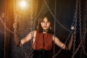 Lovely teen girl model 15-16 year posing in dark industry room, looking at camera through chains. Chic teenage girl actress in retro brown dress. Fashionable stylish image concept. Copy ad text space