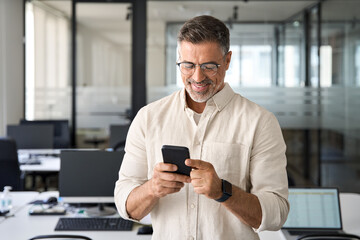 Close-up of smiling mature Latin or Indian businessman holding smartphone in office. Middle aged manager using cell phone mobile app. Digital technology application and solutions for business concept.