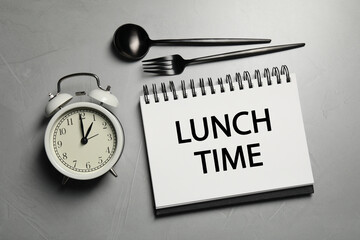 Business lunch. Notebook with phrase Lunch Time, cutlery and alarm clock on light gray table, flat...