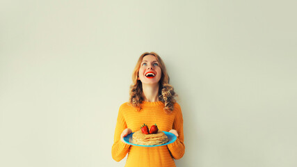 Cooking, portrait of happy laughing cheerful young woman housewife holding plate with stack of pancakes with strawberries on gray wall background