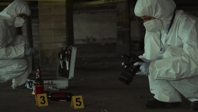 Medium full shot of two male or female forensic investigators in white overalls and masks examining urban crime scene, taking photos, collecting samples, with flashing red and blue police car lights
