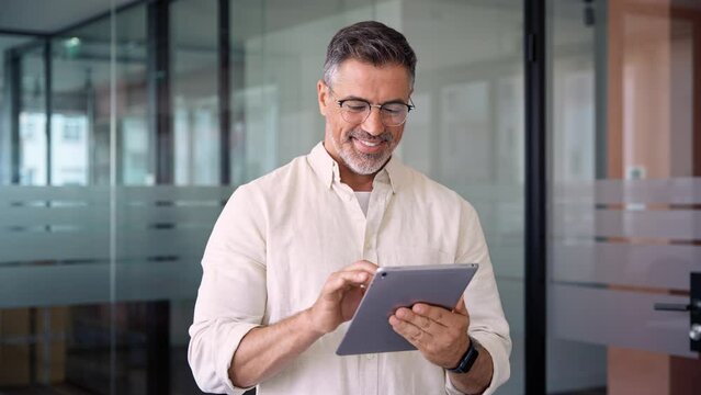 Happy Latin Hispanic bearded stylish mature adult professional business man, smiling Indian senior businessman CEO holding digital tablet using fintech tab application standing inside company office.